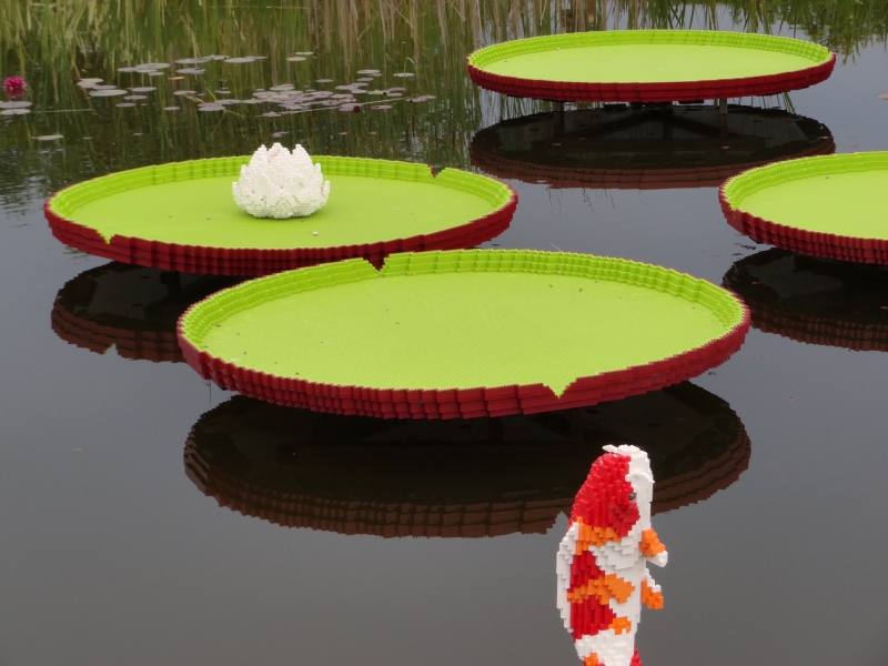 Models of large lily pads sit just above the water in a pond with a red, orange, and white LEGO fish jumping out of the water nearby