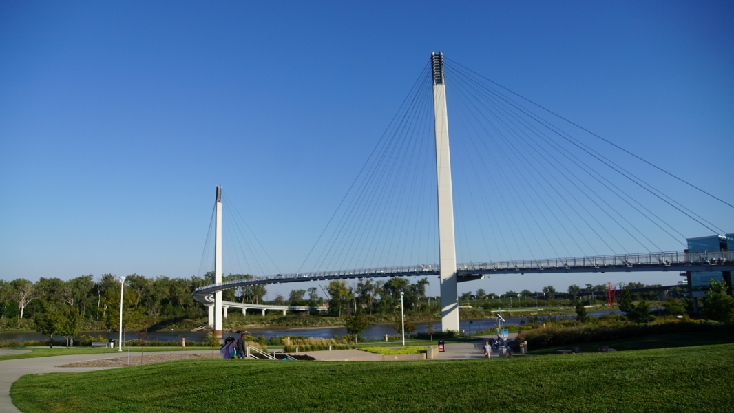 The photograph captures a modern suspension bridge with a prominent, tall white pylon supporting numerous straight, slender cables that hold up the bridge deck. The sky is clear and blue, indicating good weather conditions. Below the bridge, there is a well-maintained green park area with neatly trimmed grass and several trees scattered around. A curved footpath winds through the park, and there are a few people visible in the distance enjoying the outdoor space.