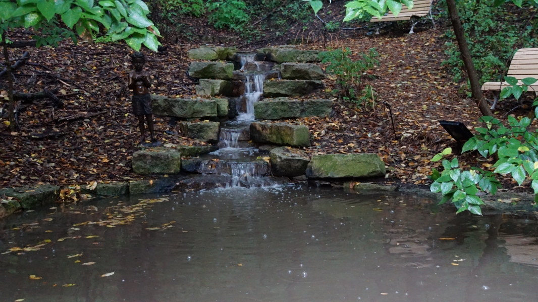 Water cascades down moss-covered rocks into a tranquil pond below. Surrounding the waterfall are green plants and trees, with some leaves showing early signs of autumnal color change. Fallen leaves are scattered across the ground, indicating early fall. A dark-colored statue stands to the left of the waterfall, blending with the natural environment. Wooden benches are partially visible on either side, suggesting a place for relaxation or contemplation in nature.