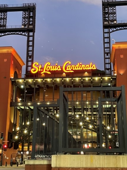 A photo of the St. Louis Cardinals stadium, Busch Stadium, at night. The iconic red brick facade and the large, illuminated St. Louis Cardinals sign are visible. The stadium's gates are closed, and there are lights on the surrounding buildings.