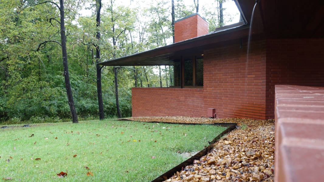 A photo of the Frank Lloyd Wright House in Ebsworth Park. The house is a low-slung, brick structure with a cantilevered roof and large windows. It is surrounded by trees and a grassy lawn. Rain runs off the roofinto a pebble filled band along one side of the house,