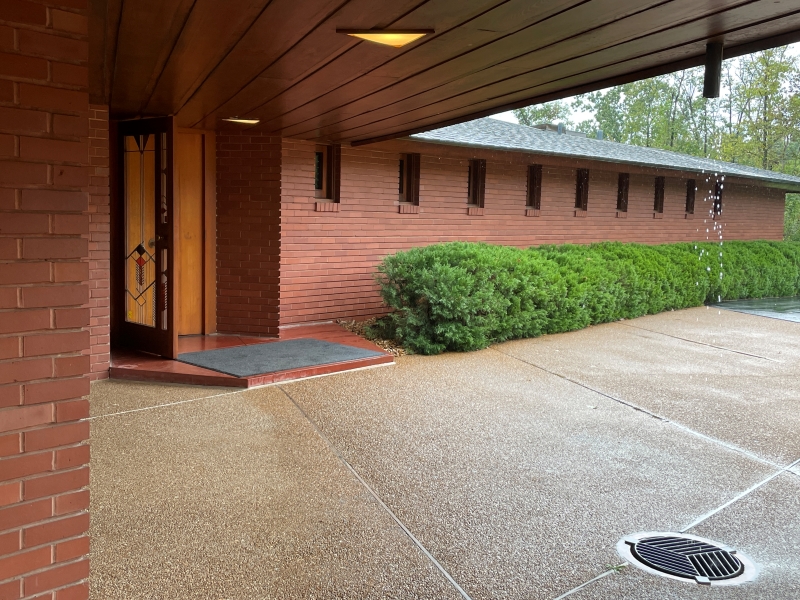 The front entrance of the Frank Lloyd Wright House in Ebsworth Park as seen from the carport. The house has a brick facade and a cantilevered roof supported by wooden beams. A wooden front door with a geometric design is visible. A row of bushes lines the side of the house, and a rain falls through a drain positioned directly over a storm drain in the driveway.