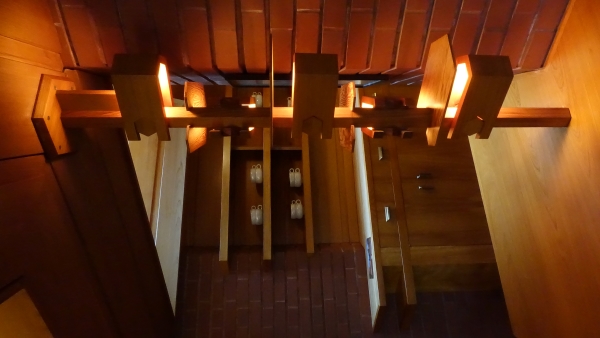 The image shows a unique wooden lamppost hanging above a table. The lamppost has several bulbs surrounded by wood shades and flat shelves positioned to defuse ligth into the space. The fixture is surrounded by brick walls and wooden cabinetry. There is a kitchen counter with dishes and utensils visible in the background.
