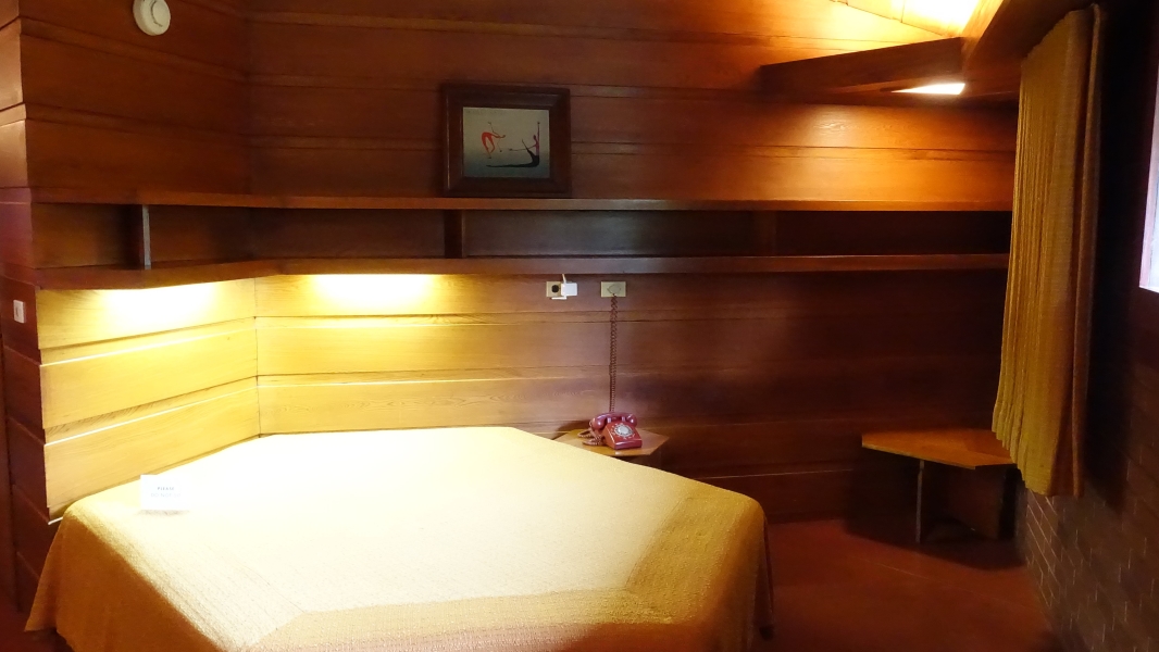 A photo of a bedroom which has a hexagonal bed with a yellow bedspread, and walls lined with warm-toned wood paneling. There is a small table with a telephone on it, and a window with curtains is visible in the background. The overall atmosphere of the room is cozy and inviting.