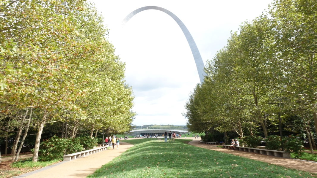 A photo of the Gateway Arch National Park in St. Louis, Missouri. The iconic Gateway Arch, a tall, stainless steel monument shaped like a soaring arch, dominates the center of the image. The arch is surrounded by a park with trees, grass, and a paved walkway. People can be seen walking along the walkway and enjoying the view.