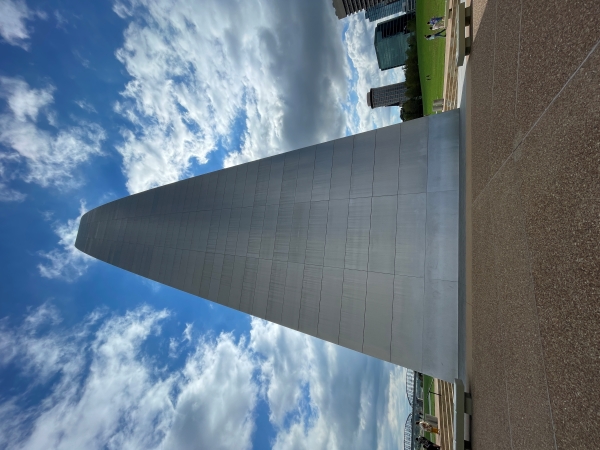 A photo of the Gateway Arch in St. Louis, Missouri, from a low angle. The arch's massive, stainless steel structure towers over the viewer, reaching towards a bright blue sky with fluffy clouds. The base of the arch is visible, with a concrete plaza and a few people walking around.