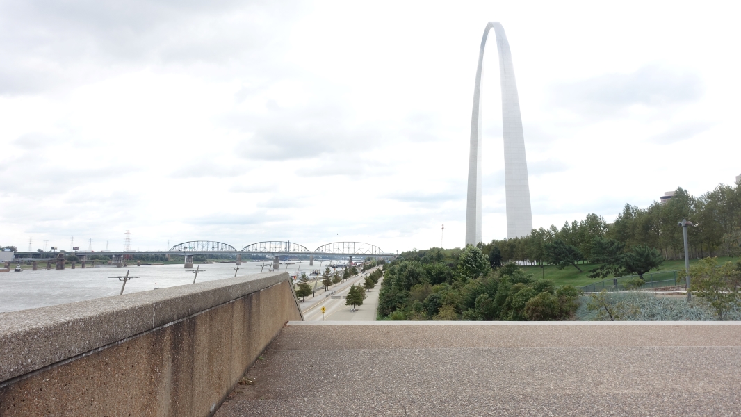 The iconic Gateway Arch stands tall in the background, towering over the Mississippi River. A bridge with multiple spans crosses the river in the foreground, and there are trees and greenery along the riverbank.