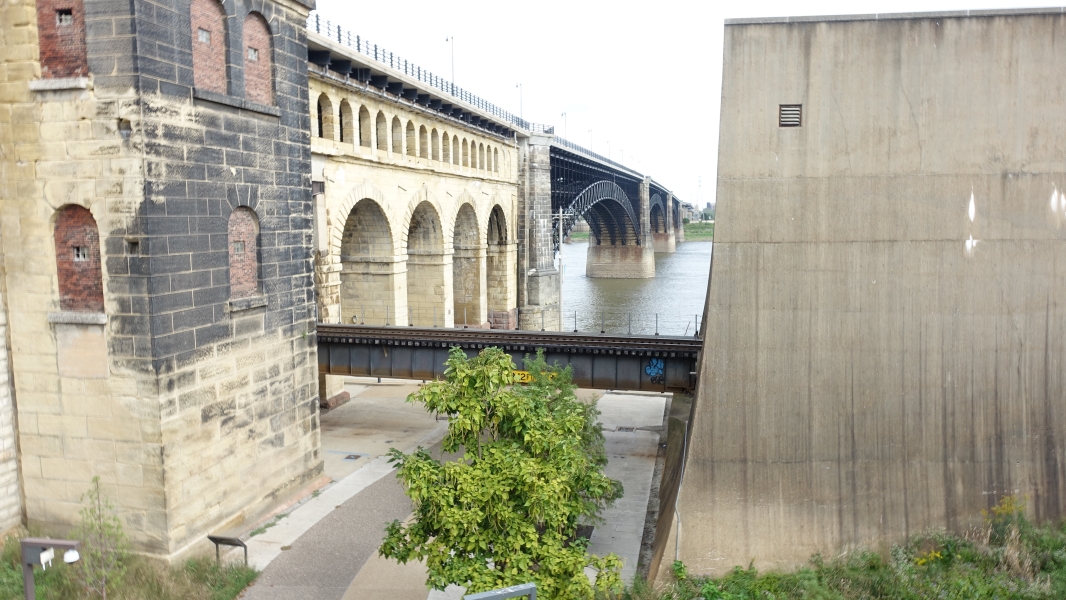 A photo of a historic stone bridge with multiple arches spanning a river. The bridge is flanked by a tall, concrete wall on one side and a brick building on the other. A small tree grows in a space between the bridge and the building, and there is a paved walkway leading towards the river. The sky is cloudy, and the water in the river is calm.