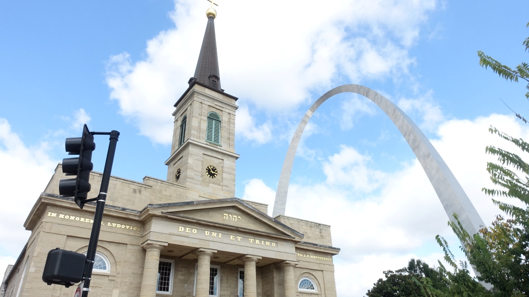 A photo of the Basilica of Saint Louis, King of France, and the Gateway Arch in St. Louis, Missouri. The basilica is a large, historic church with a tall steeple and a clock tower. The Gateway Arch, a towering stainless steel monument, stands in the background. The sky is blue with fluffy white clouds.