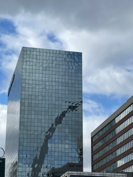 A photo of a tall, modern skyscraper with a glass facade reflecting the Gateway Arch. The building is surrounded by patches of blue sky between gray clouds.